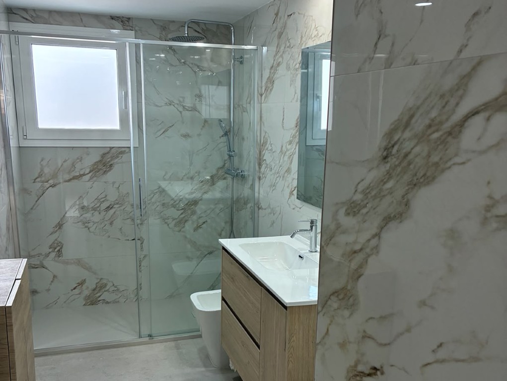 Baño reformado con ducha de vidrio, mueble de madera y revestimiento efecto mármol