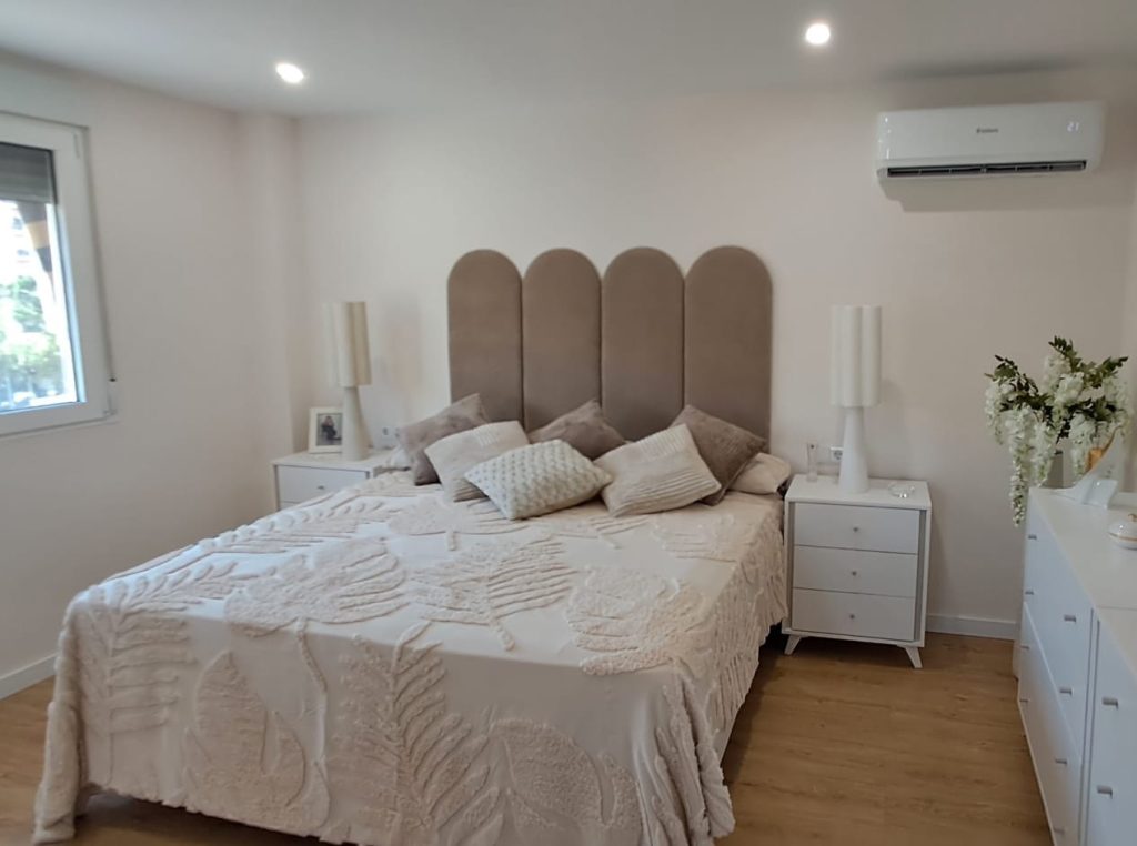Dormitorio reformado con cabecero tapizado, mesillas blancas y suelo de madera